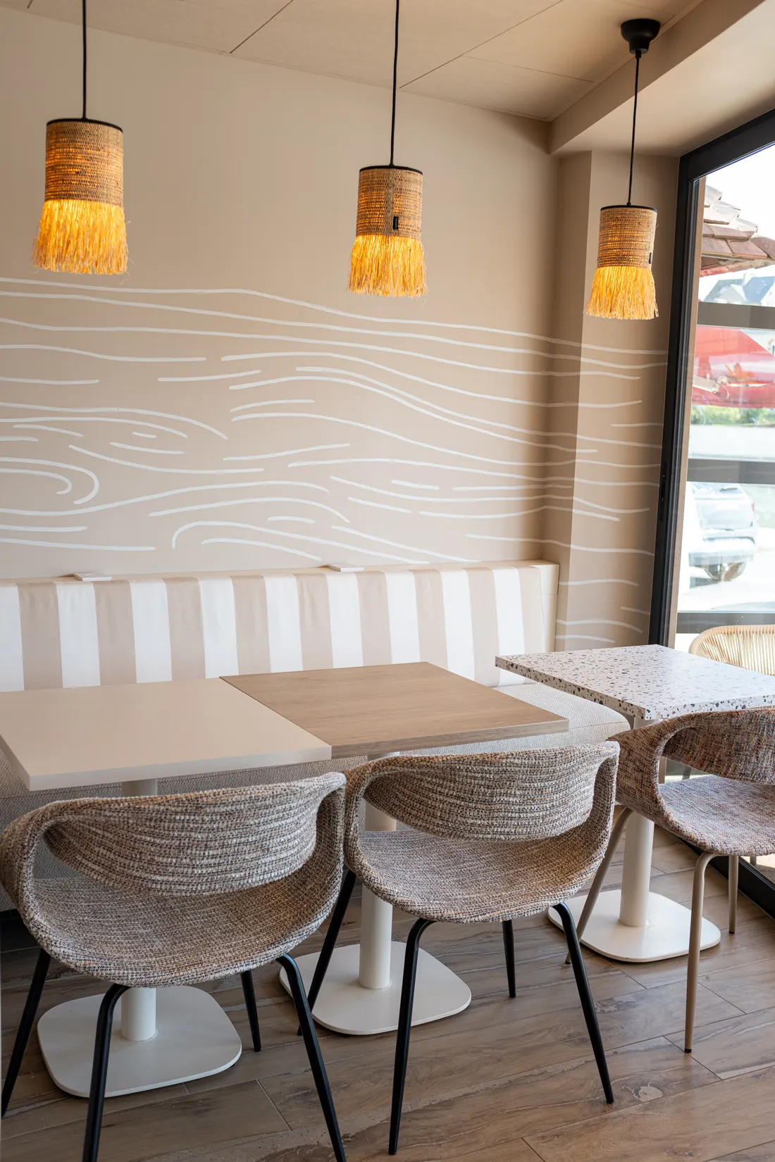 Salle du restaurant : banquettes rayées beige et blanc, tables en bois clair, suspensions en raphia, mur aux lignes ondulées