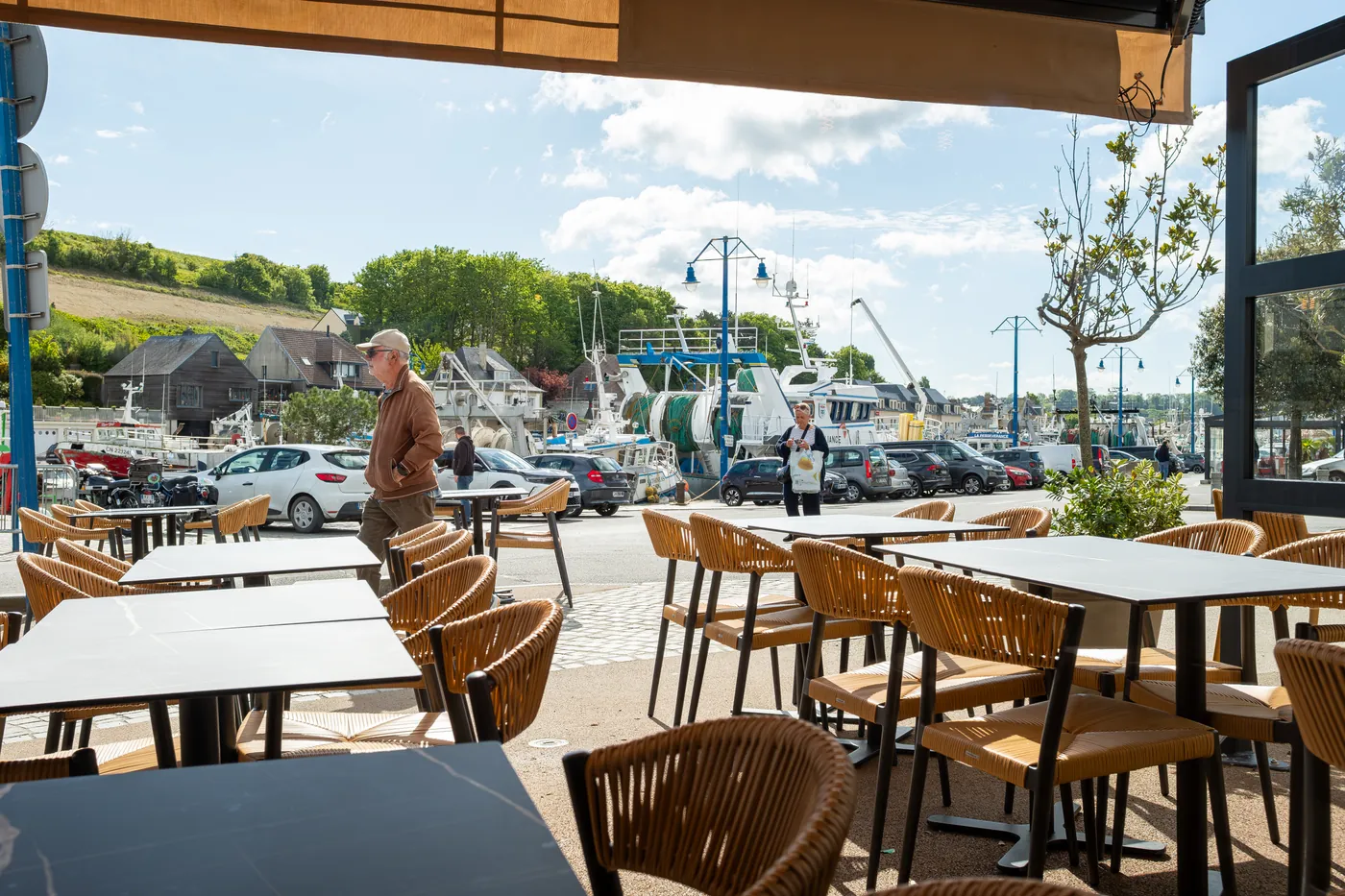 Terrasse aux chaises en rotin face au port de Port-en-Bessin, bateaux de pêche à quai en arrière-plan
