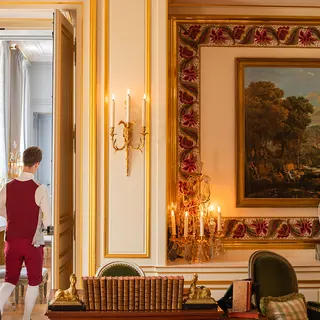 Salon d'hôtel aux boiseries dorées, un valet en livrée rouge de dos, photo pour Miele