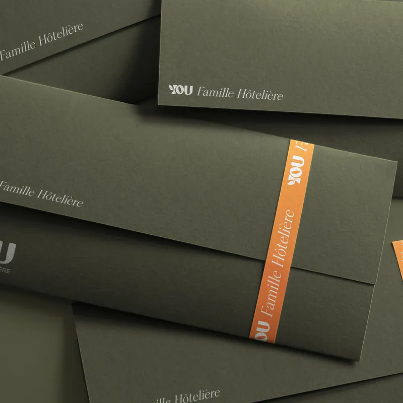 Enveloppes vert olive au logo YOU Famille Hôtelière, l'une fermée par une bande orange