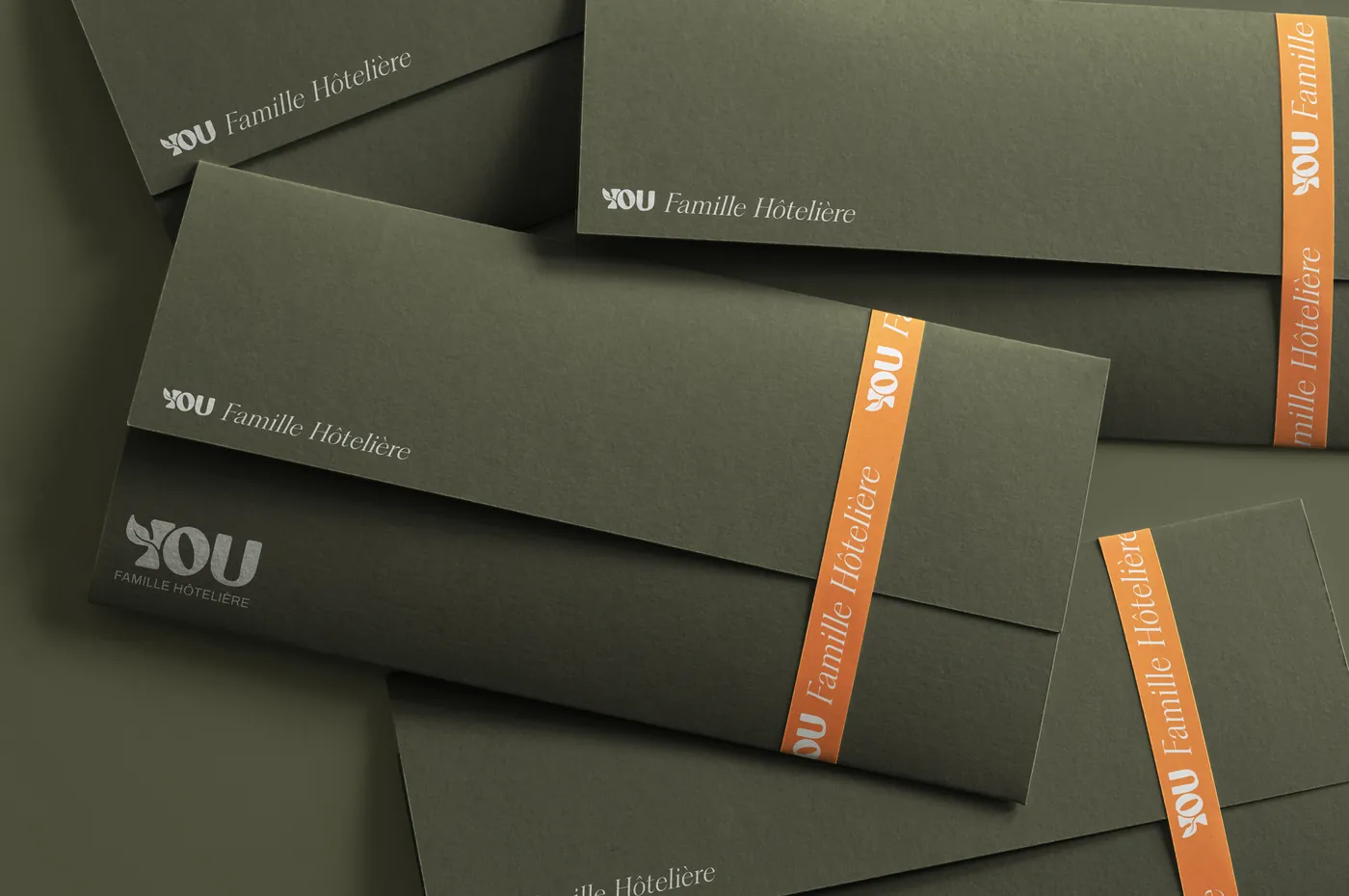 Enveloppes longues vert olive au logo YOU, cerclées d'une bande orange « Famille Hôtelière » en lettres blanches