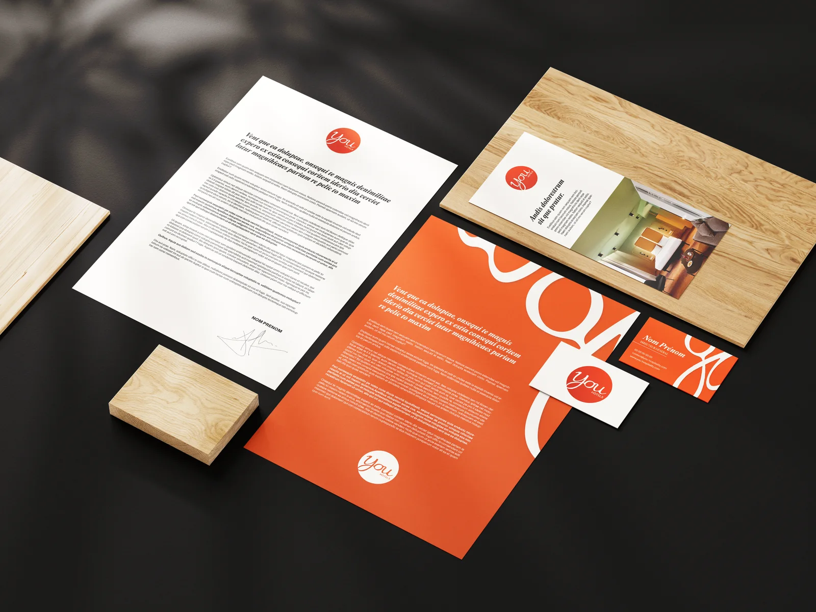 Papeterie YOU Hotels sur fond noir : lettre, cartes et chemise orange aux grandes boucles blanches du « You »