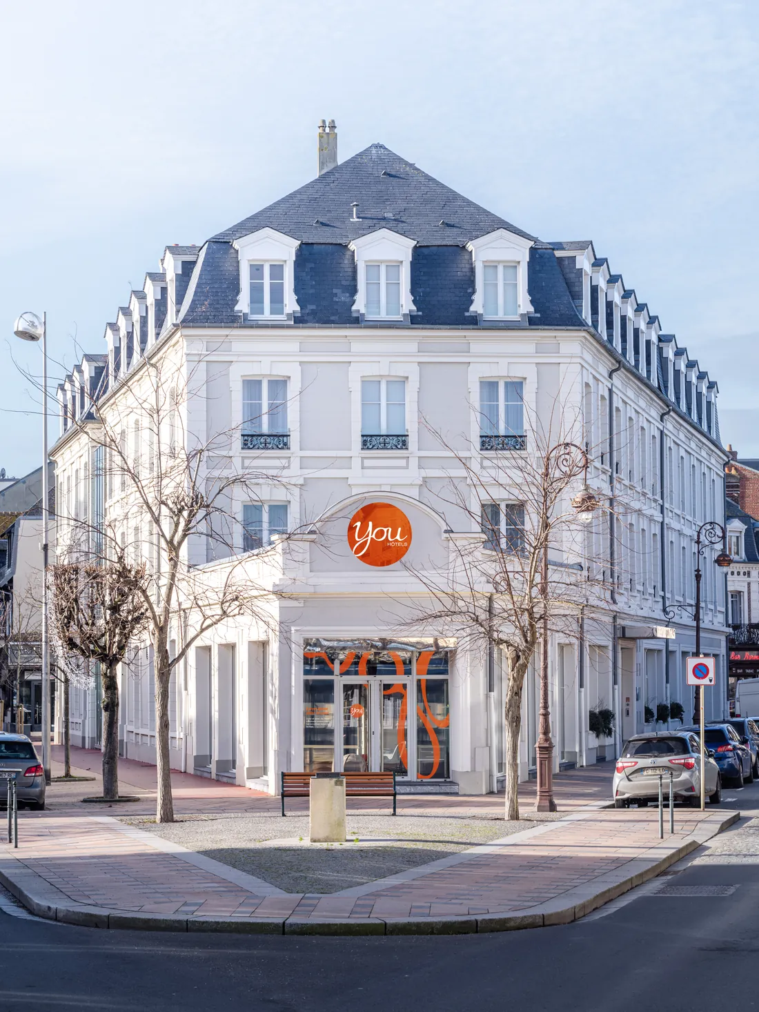 Simulation de façade de l'hôtel YOU Deauville : immeuble blanc d'angle, enseigne ronde orange « You » au-dessus de l'entrée